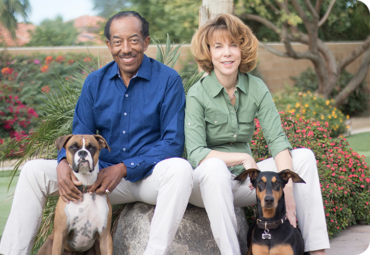 A man in a blue shirt and white pants sits on a rock beside a woman in a sage green shirt and white pants with a Doberman Pinscher and a Boxer with them and floral shrubs in the background